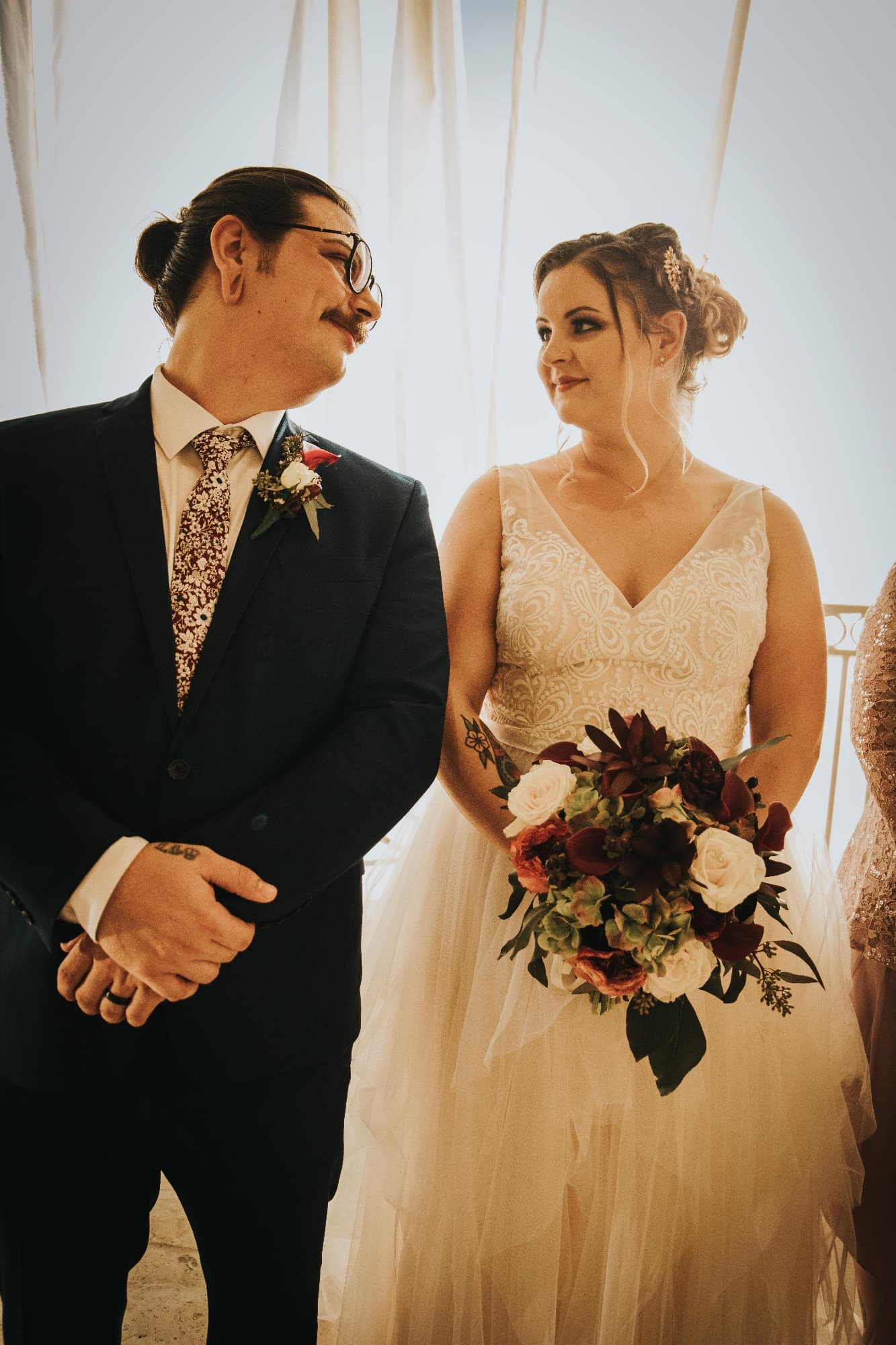 A groom in a navy suit and floral tie smiles sideways at a bride holding a burgundy and blush bouquet, in bright backlight.