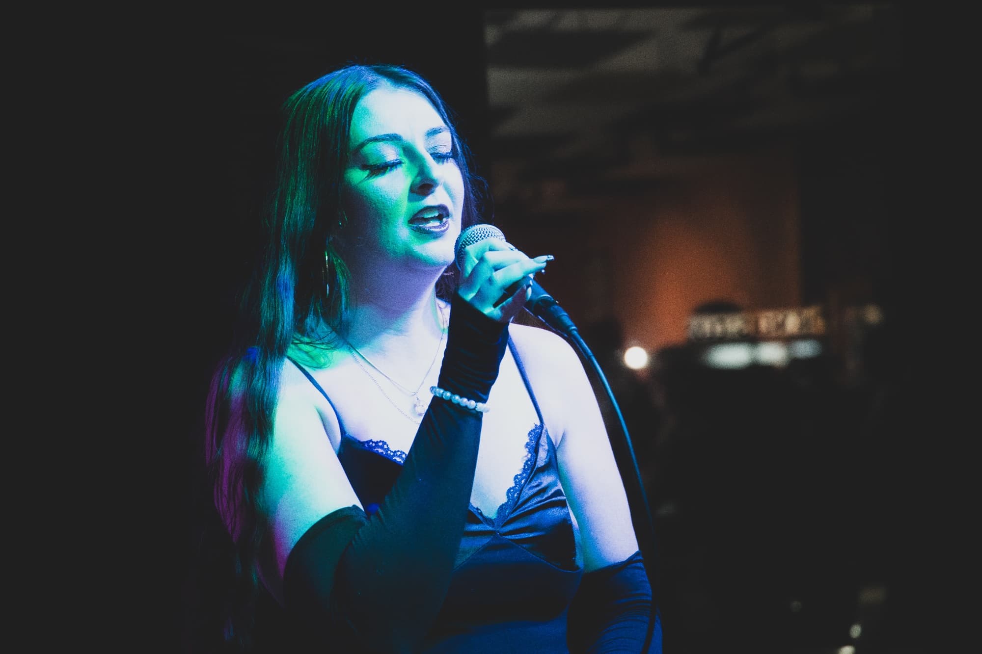A woman with long dark hair sings into a microphone with her eyes closed, lit in teal and blue stage light.
