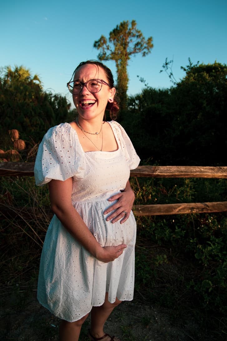 A laughing woman in glasses and a white eyelet dress cradles her belly beside a wooden fence in warm evening light.