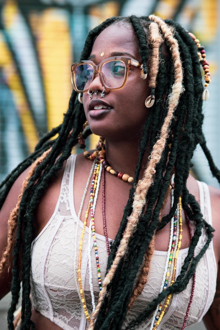 A woman in oversized amber glasses looks off to the side, wearing long dark locs with blonde strands, layered beaded necklaces and a lace top, in front of a colorful mural.