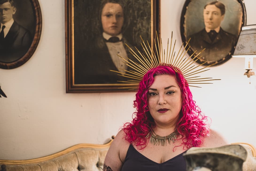 A woman with bright pink curly hair and a gold spiked halo crown sits on a vintage sofa before antique framed portraits.