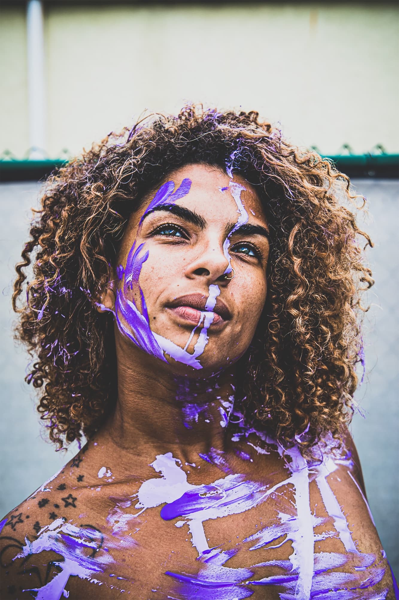 Close portrait of a woman with curly hair and a nose ring, gazing upward, her face and shoulders streaked with purple and white paint.