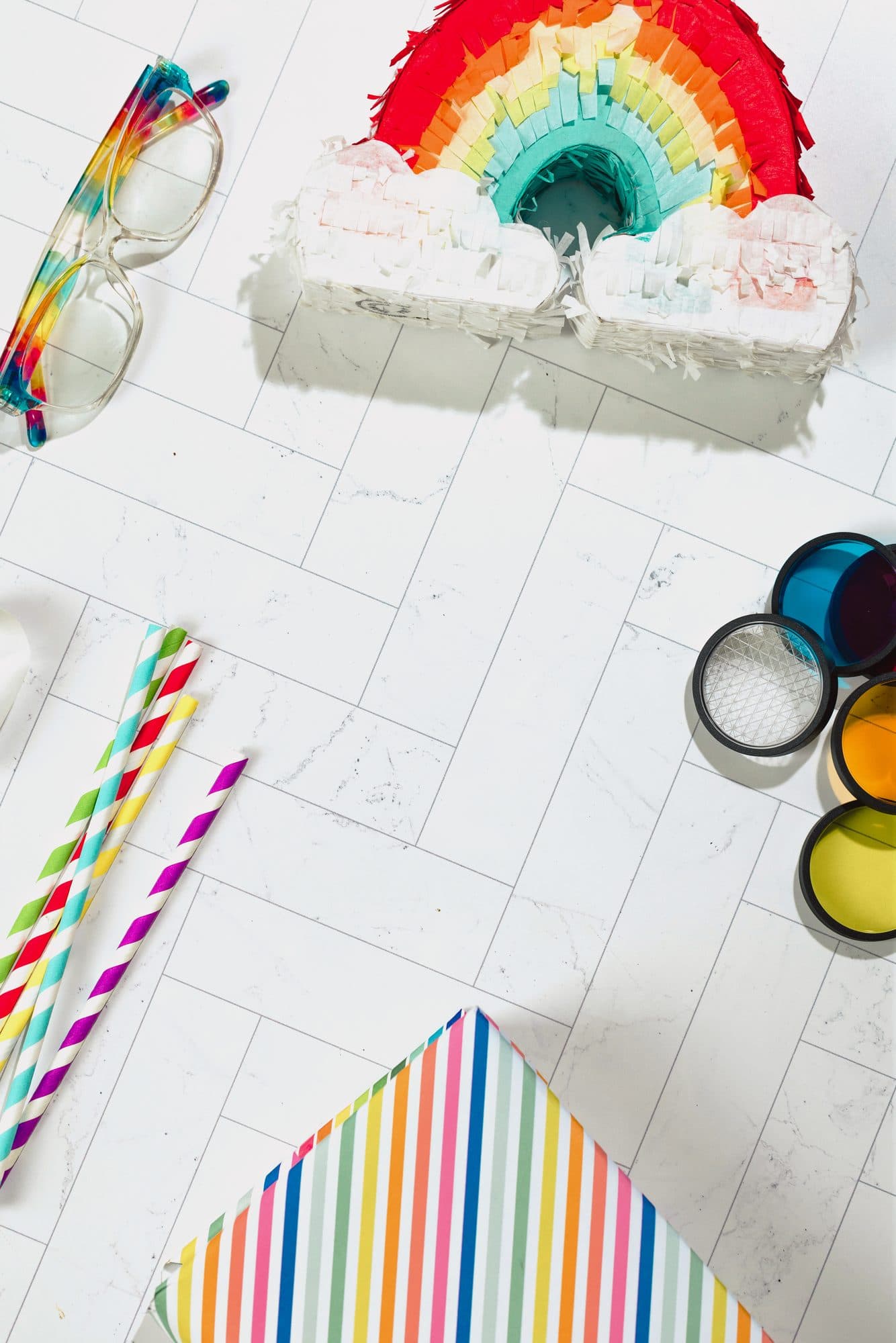 Flat-lay on white marble: rainbow piñata, striped paper straws, colored lens filters