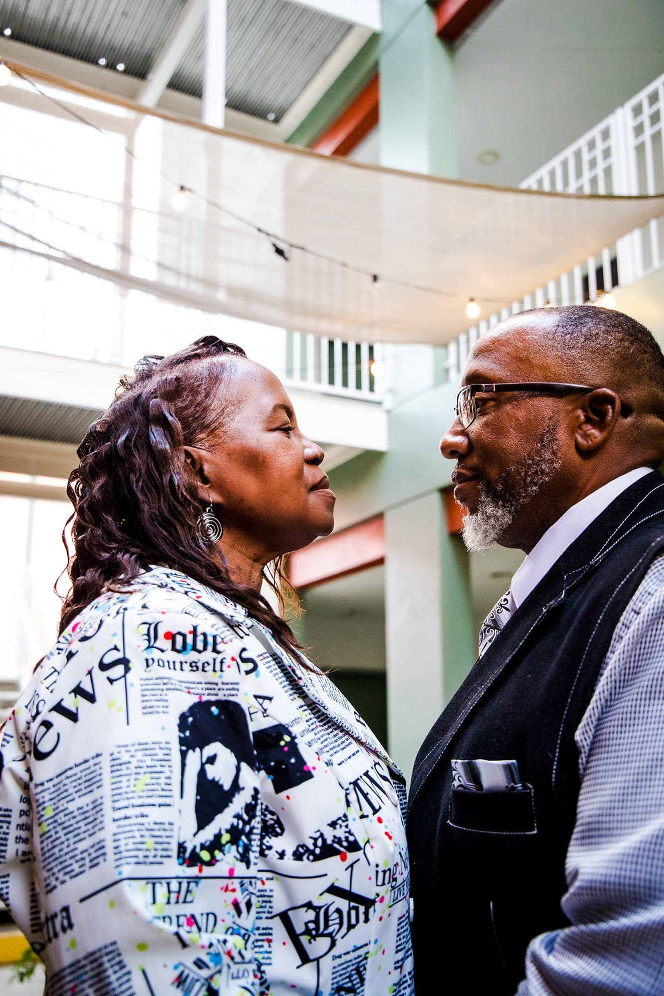 A couple nose to nose in a bright atrium, she in a newsprint jacket