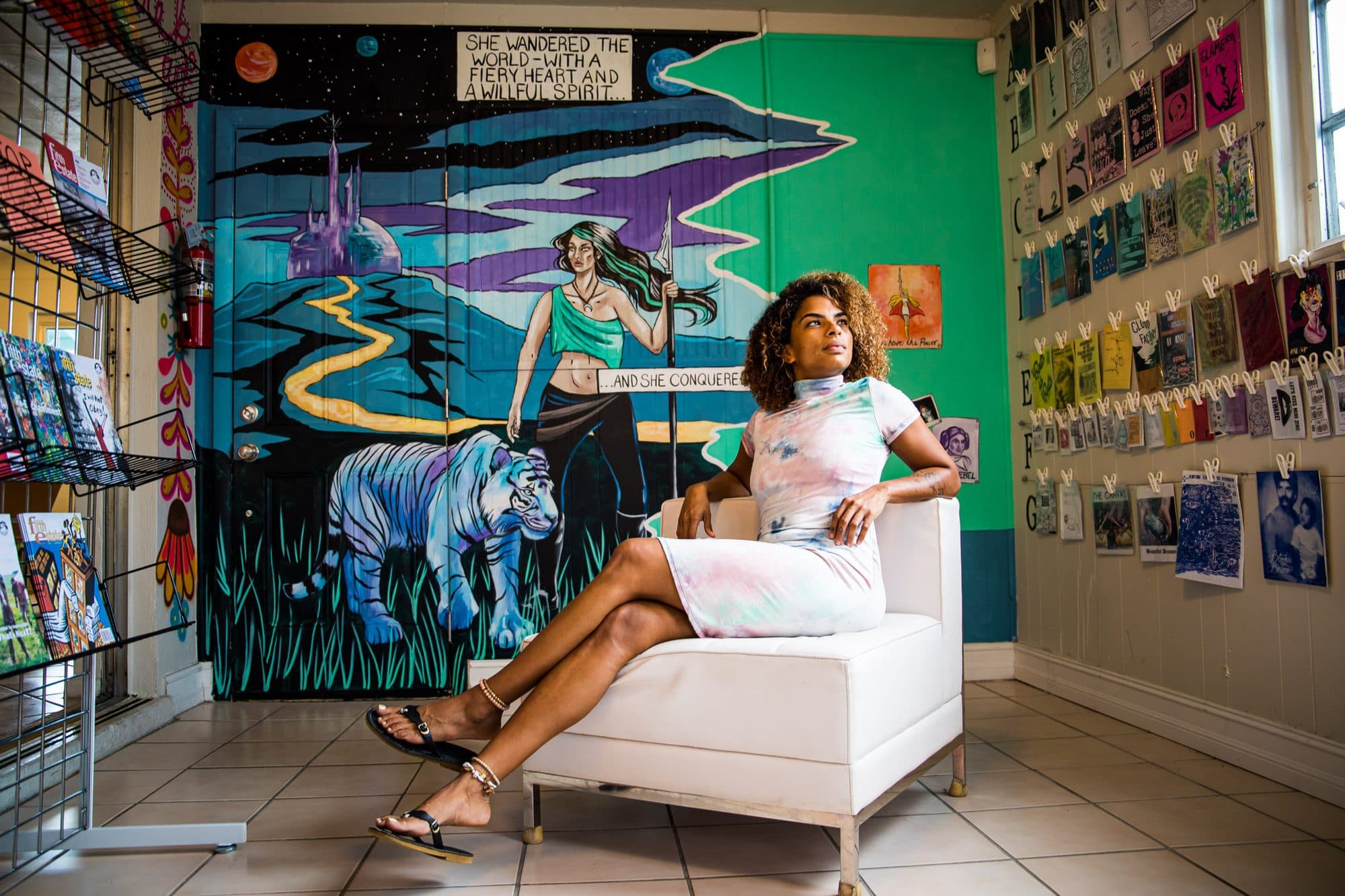 Editorial portrait: a woman in a tie-dye dress seated on a white chair beneath a tiger mural in a gallery shop