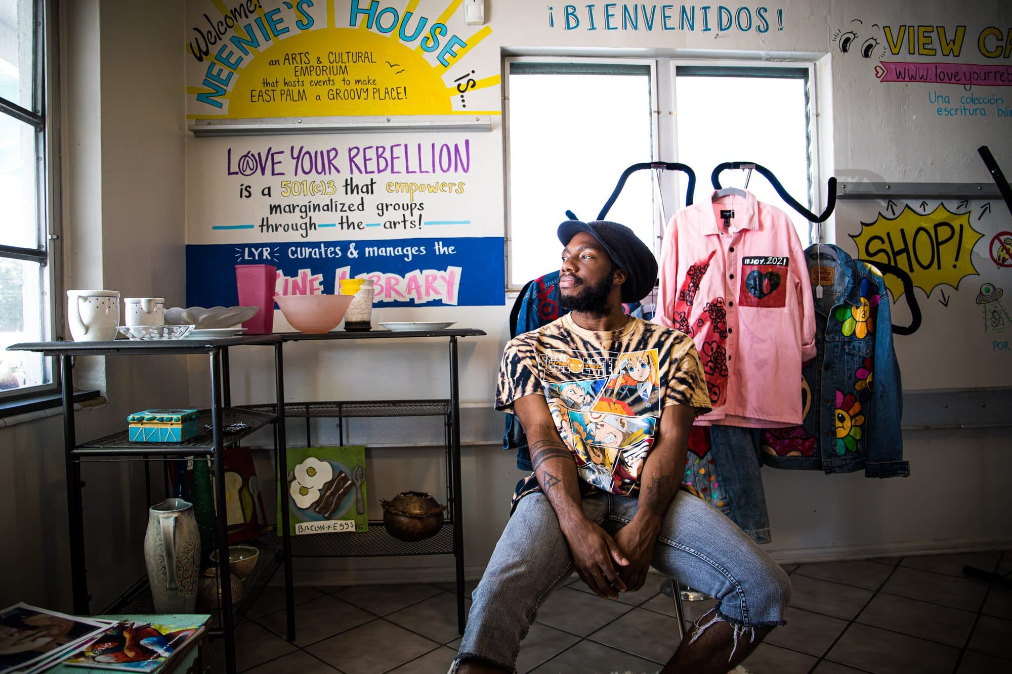 A man in tie-dye seated in his shop beneath a hand-lettered welcome sign