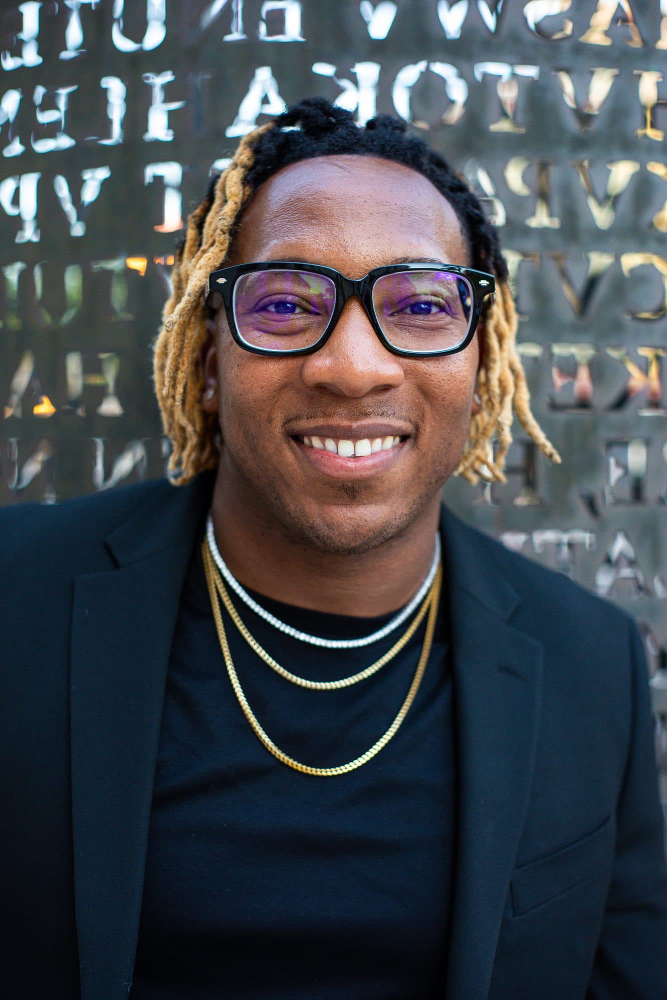 A man in glasses and a black suit in front of a wall of cut-metal letters