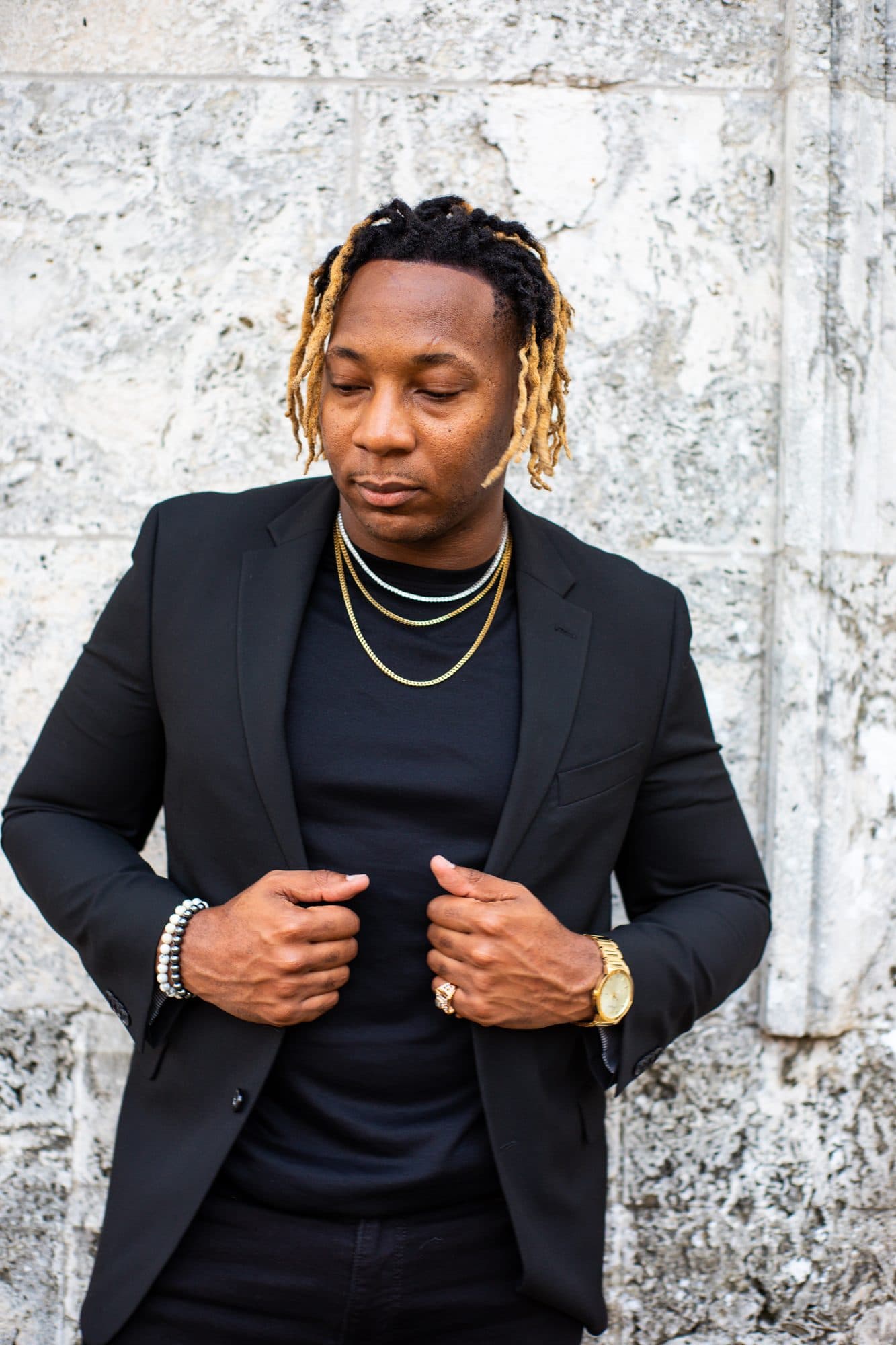 Editorial portrait of a man in a black suit and gold chains adjusting his jacket against a coral stone wall