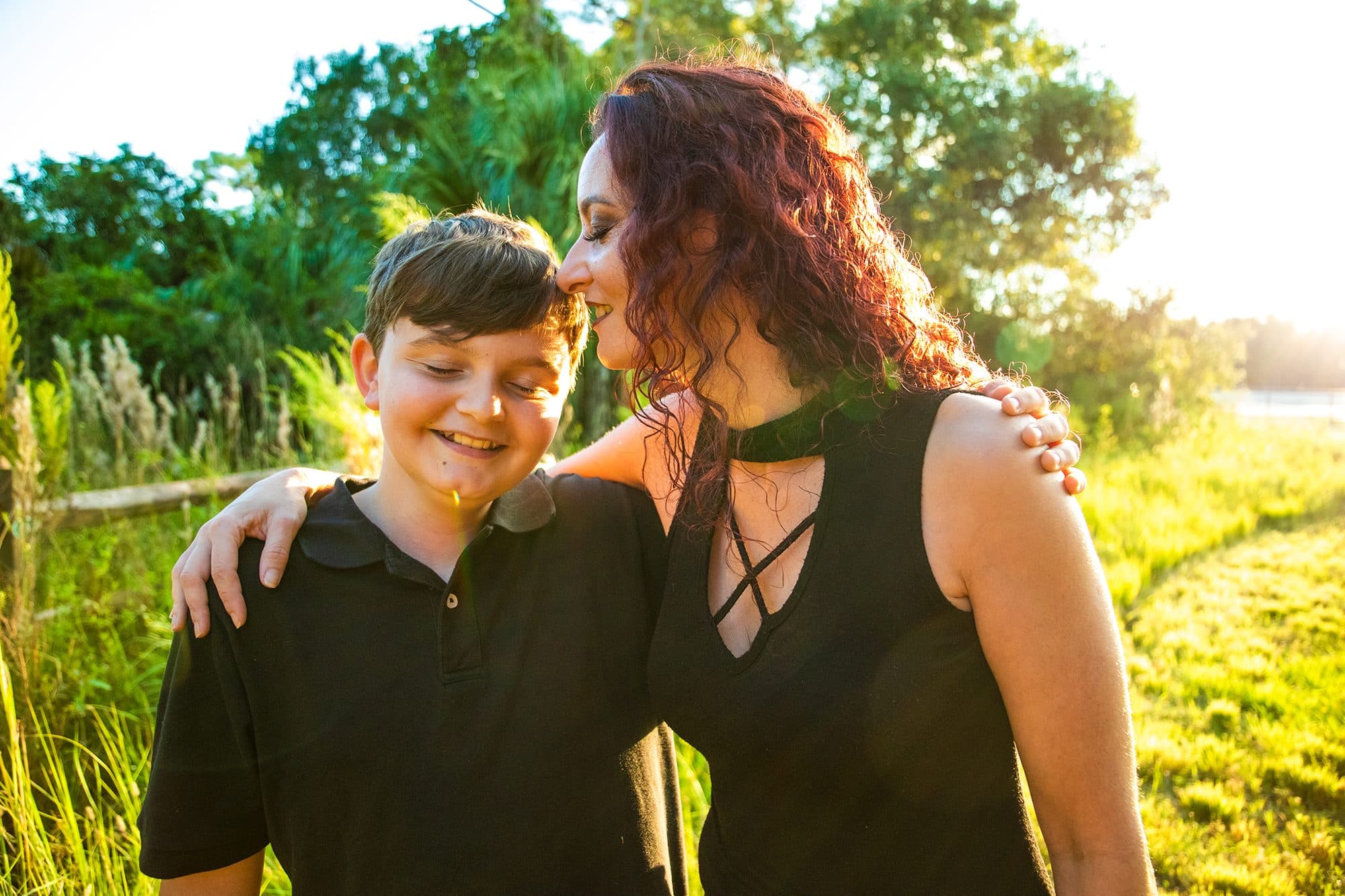 Mother pressing a kiss to her son's head in tall golden grass at sunset