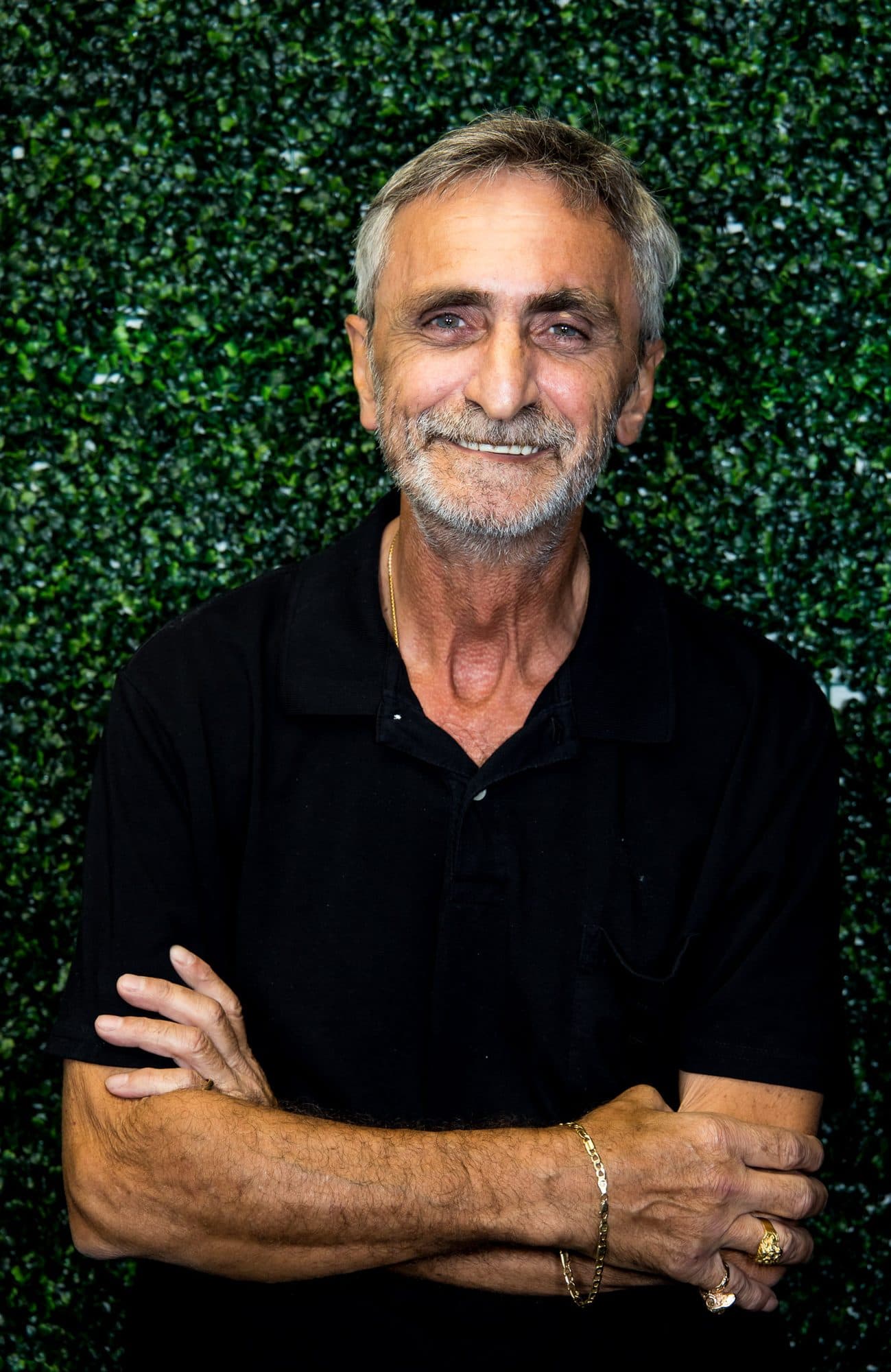 Portrait of a silver-haired man in a black polo, arms crossed, against a greenery wall
