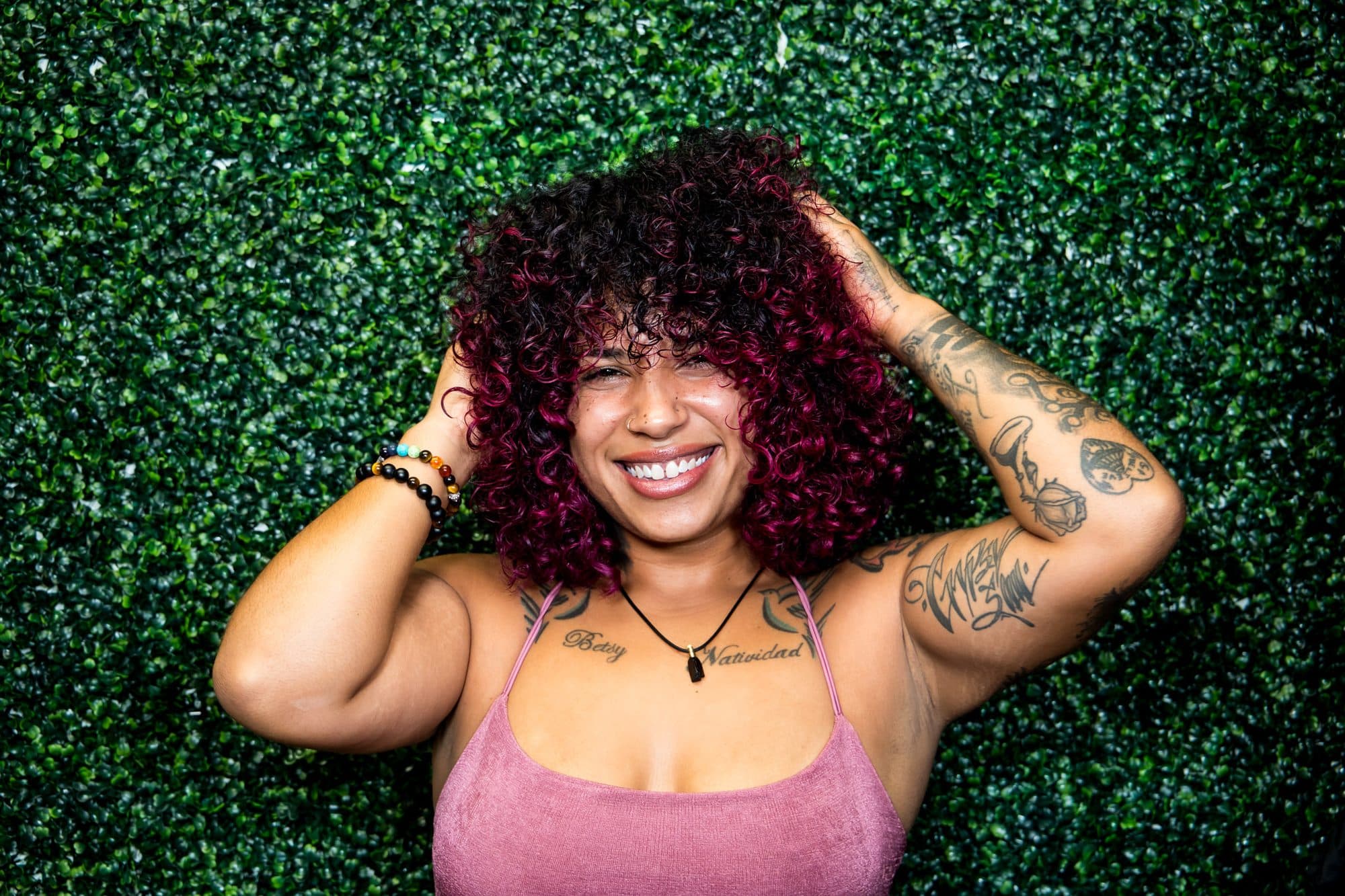 Portrait of a woman with red curls and tattoos, hands in her hair, against a greenery wall
