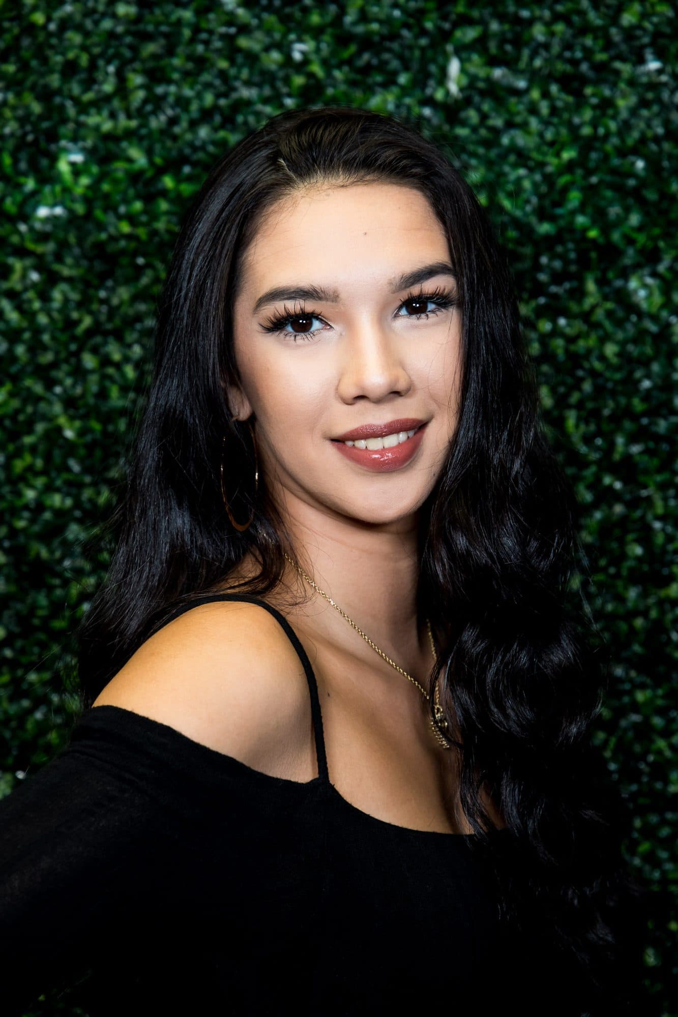 Portrait of a woman with long dark hair and an off-shoulder top against a greenery wall