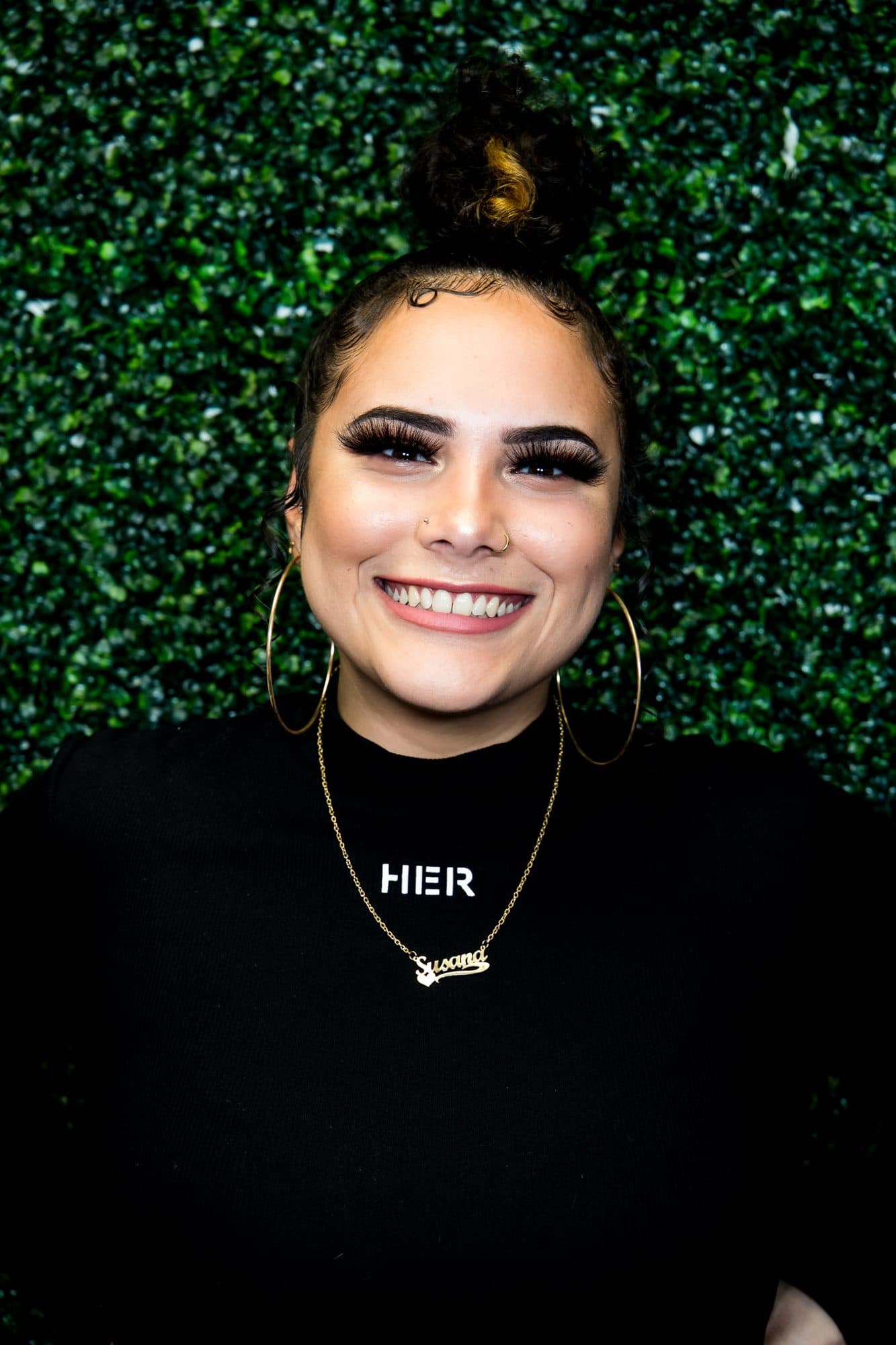 A smiling woman with a top-knot and a necklace that reads HER against a greenery wall