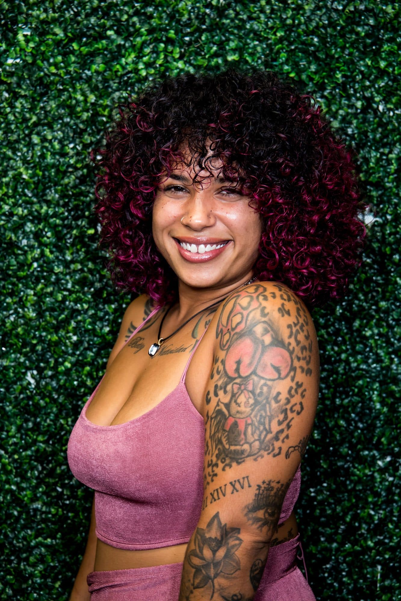 A laughing woman in a pink top with arm tattoos against a greenery wall