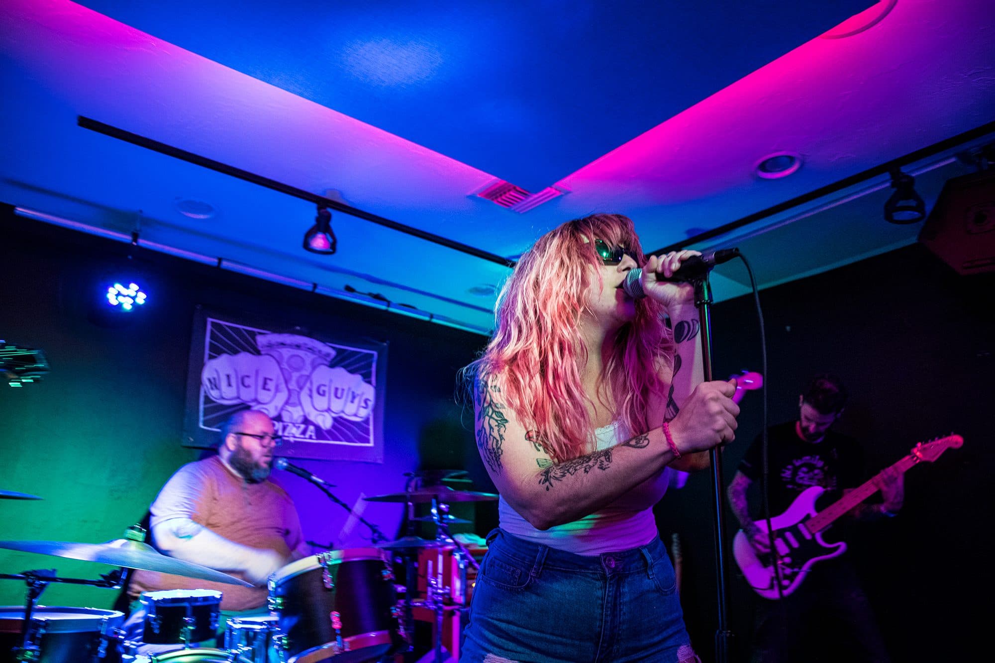 Singer with pink hair mid-note, drummer lit blue behind her