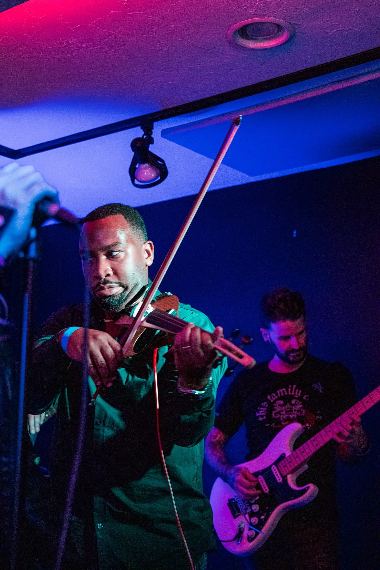 A violinist lit purple, guitarist behind him in the shadows