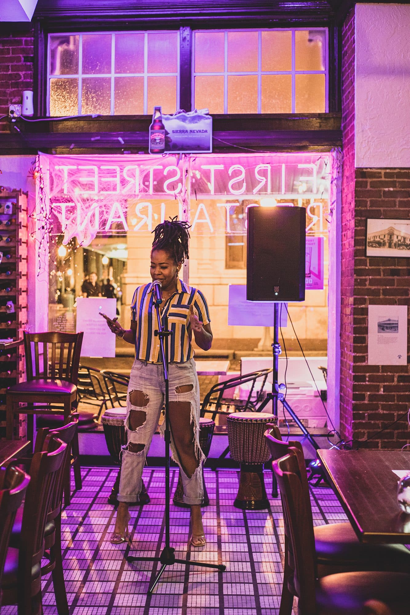 A performer on stage beneath a neon First Street sign, pink light spilling across the room
