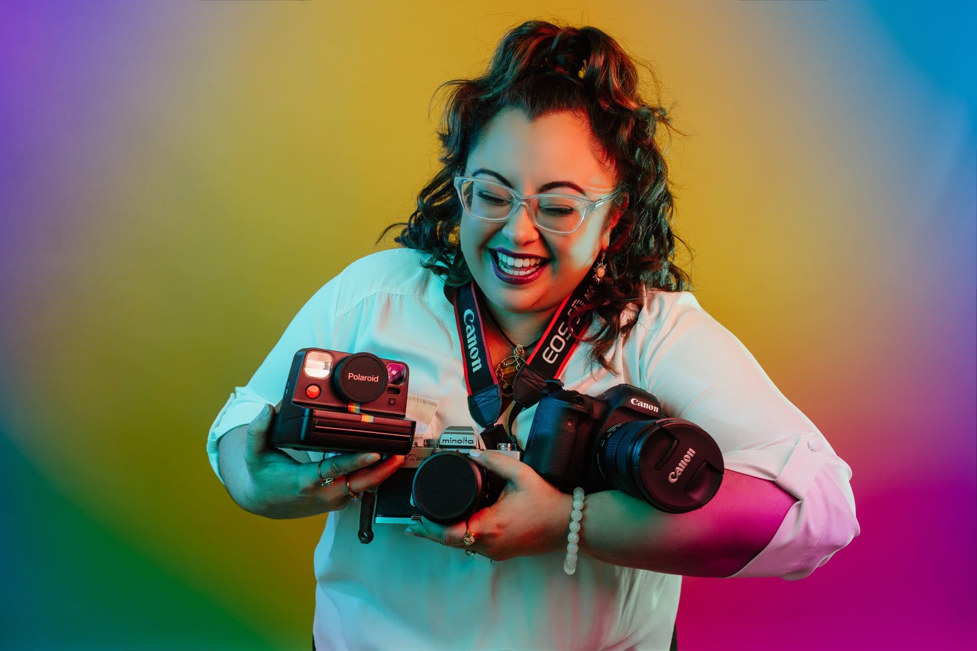 Tylor Hooks laughing while hugging an armful of cameras — a Polaroid, a Minolta and a Canon — in front of a rainbow gradient
